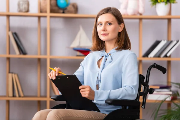 Psychologue féminine positive avec presse-papiers regardant la caméra en fauteuil roulant sur fond flou — Photo de stock