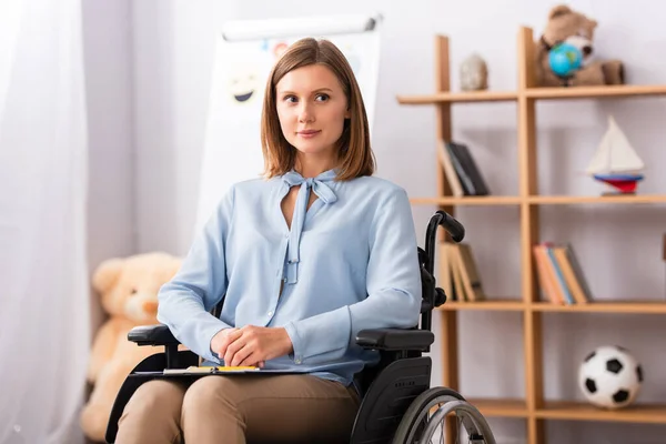 Psicólogo positivo com as mãos apertadas olhando para longe enquanto sentado em cadeira de rodas com escritório borrado no fundo — Fotografia de Stock