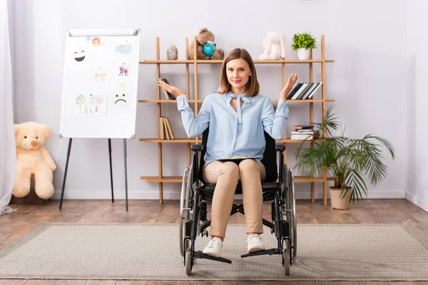 Piena lunghezza di psicologo positivo con gesto scrollata di spalle seduto in sedia a rotelle in ufficio — Foto stock