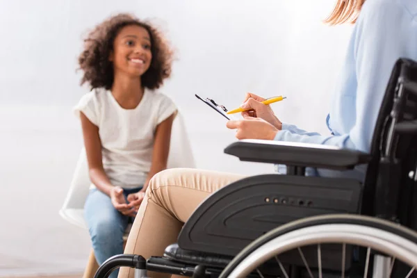 Psychologist writing on clipboard while sitting in wheelchair with blurred happy african american girl on background — Stock Photo