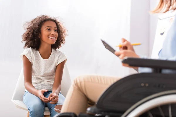 Heureuse fille afro-américaine assise sur une chaise avec psychologue floue en fauteuil roulant au premier plan — Photo de stock