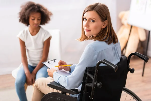 Psychologue positif écrit sur presse-papiers tout en étant assis en fauteuil roulant avec une fille afro-américaine floue sur fond — Photo de stock