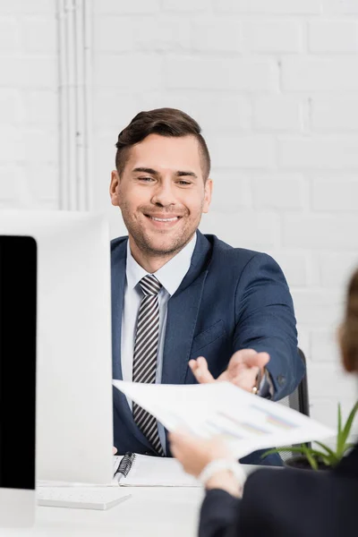 Feliz hombre de negocios tomando documento con gráficos, mientras está sentado en el lugar de trabajo con monitores de computadora en primer plano borroso - foto de stock