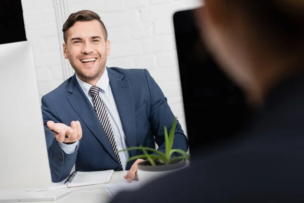 Happy executive gesturing, while sitting at workplace with blurred female co-worker on foreground — Stock Photo