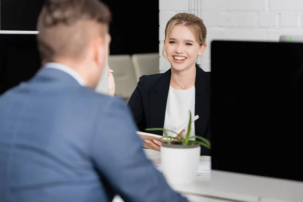 Femme d'affaires blonde riant, tout en étant assis sur le lieu de travail avec un collègue flou au premier plan — Photo de stock