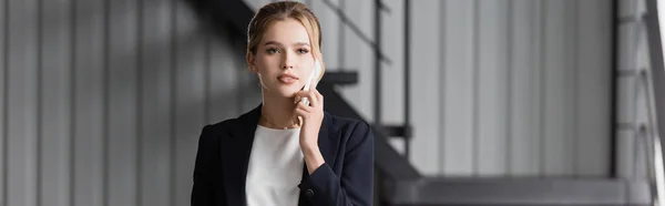 Mujer de negocios rubia mirando hacia otro lado, mientras habla en el teléfono móvil en la oficina sobre fondo borroso, pancarta - foto de stock