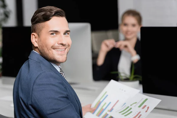 Hombre de negocios sonriente con hojas de papel mirando a la cámara, mientras está sentado en el lugar de trabajo con un compañero de trabajo borroso en el fondo - foto de stock