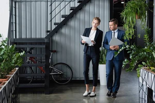 Бизнесмен с ноутбуком, глядя на бумажные листы в руках коллеги-женщины, стоя на работе — стоковое фото