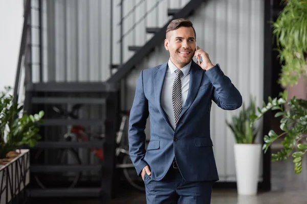 Hombre de negocios sonriente con la mano en el bolsillo mirando hacia otro lado, mientras habla en el teléfono móvil en un fondo borroso - foto de stock