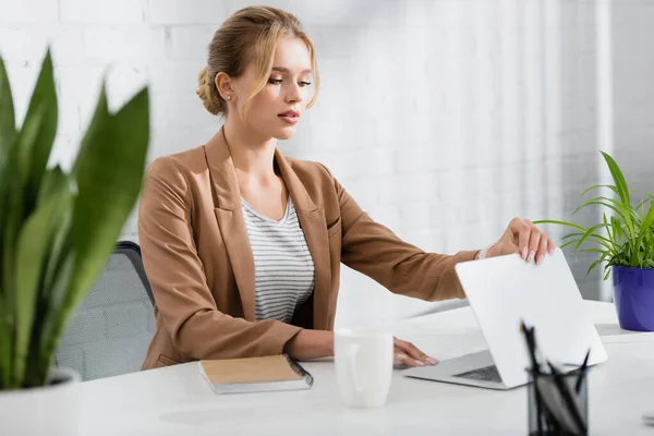 Femme d'affaires blonde fermeture ordinateur portable, tout en étant assis à la table au bureau sur le premier plan flou — Photo de stock