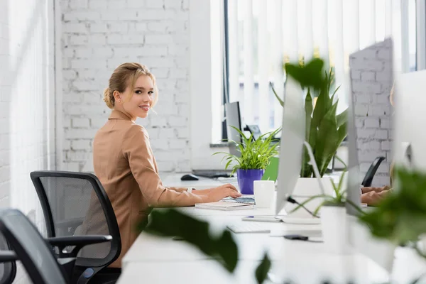 Donna d'affari sorridente che guarda la fotocamera, mentre digita sulla tastiera del computer sul posto di lavoro in primo piano sfocato — Foto stock