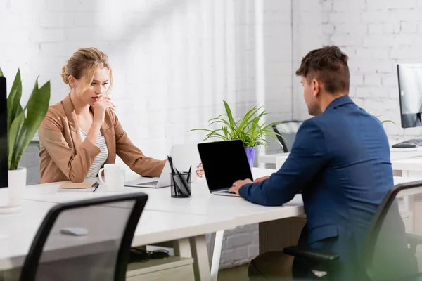 Thoughtful businesswoman looking at laptop, while sitting near colleague at workplace in office — Stock Photo