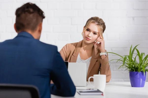 Thoughtful businesswoman looking at colleague, while sitting at table with laptops on blurred foreground — Stock Photo