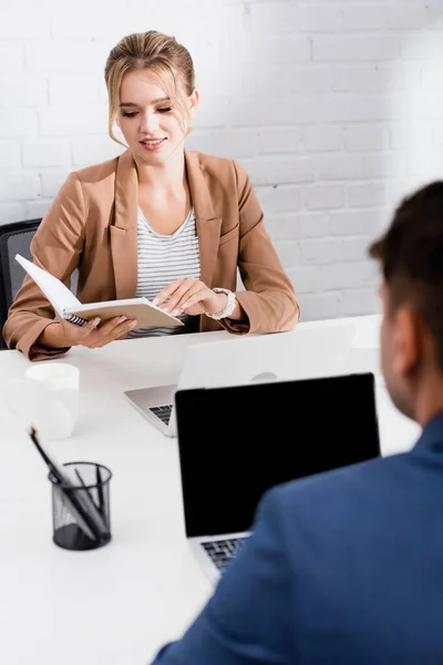 Mujer de negocios sonriente mirando el cuaderno, mientras está sentada en la mesa con computadoras portátiles en la oficina en primer plano borroso - foto de stock