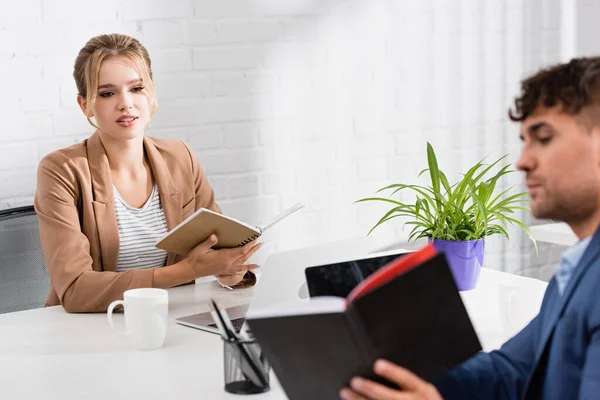 Mujer de negocios mirando el cuaderno en manos de un colega, mientras está sentado en el lugar de trabajo en primer plano borrosa - foto de stock