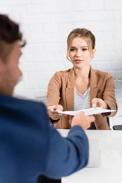 Mujer de negocios rubia tomando documento de su colega, mientras se sienta a la mesa en primer plano borrosa - foto de stock