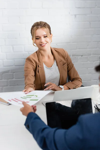 Femme d'affaires souriante donnant une feuille de papier avec des graphiques à un collègue, tout en étant assis sur le lieu de travail sur le premier plan flou — Photo de stock