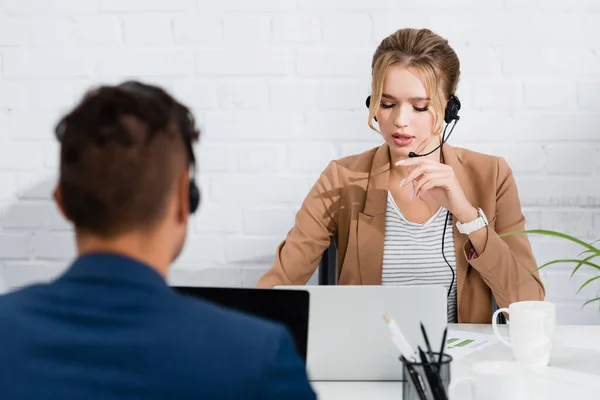 Operadora femenina en auriculares hablando, mientras está sentada en el lugar de trabajo con computadoras portátiles en la oficina en primer plano borroso - foto de stock