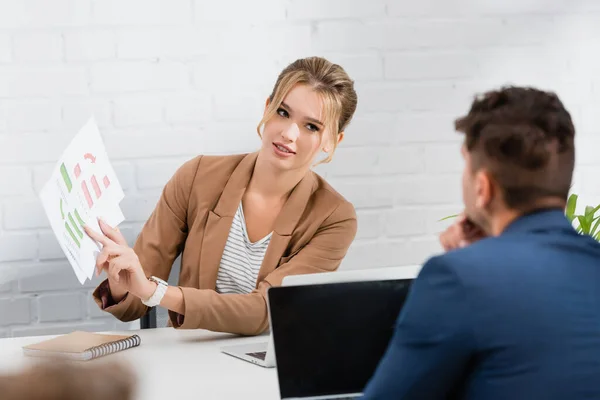 Businesswoman pointing with finger at paper sheet with charts, looking at colleague sitting at workplace on blurred foreground — Stock Photo