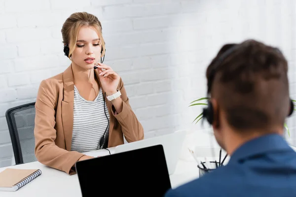 Ernste Frau im Kopfhörersprech, am Tisch sitzend mit Laptop und verschwommenem Kollegen im Vordergrund — Stockfoto