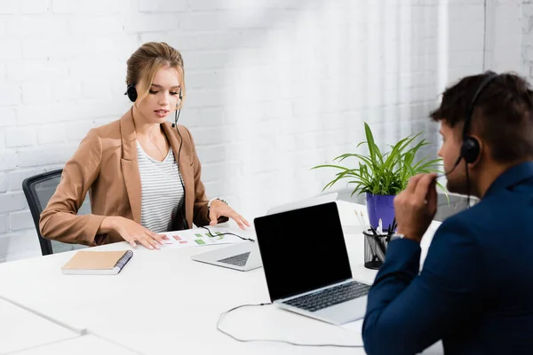 Female operator in headset talking, while looking at charts on sheet of paper at work on blurred foreground — Stock Photo