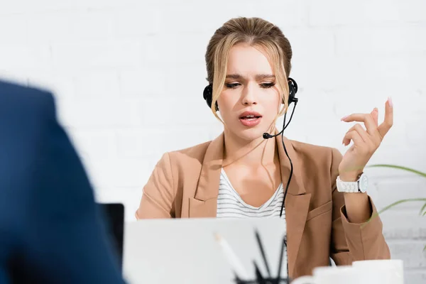 Serious woman in headset gesturing, while sitting near laptop at workplace on blurred foreground — Stock Photo