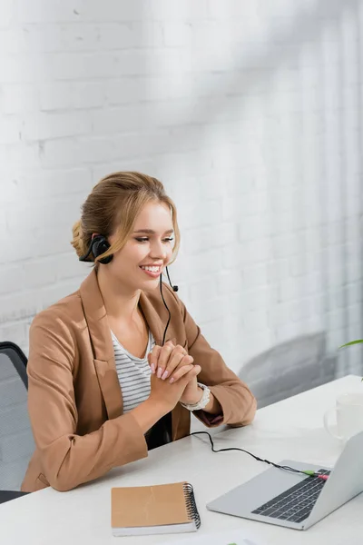 Fröhliche Beraterin mit Headset, die am Arbeitsplatz auf den Laptop schaut — Stockfoto