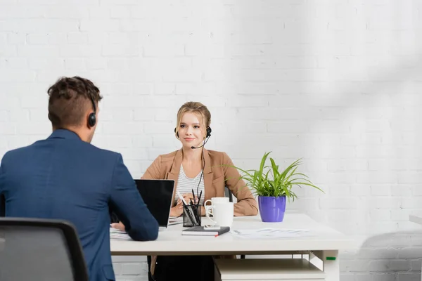 Opérateur féminin positif dans casque regardant collègue, tout en étant assis sur le lieu de travail avec des appareils numériques — Photo de stock
