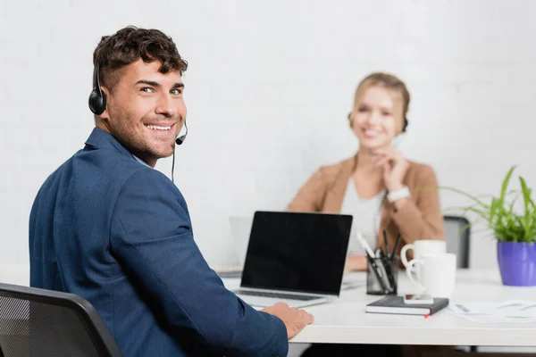 Callcenter-Bediener mit Headset blickt auf Kamera, während er am Arbeitsplatz mit digitalen Geräten auf verschwommenem Hintergrund sitzt — Stockfoto