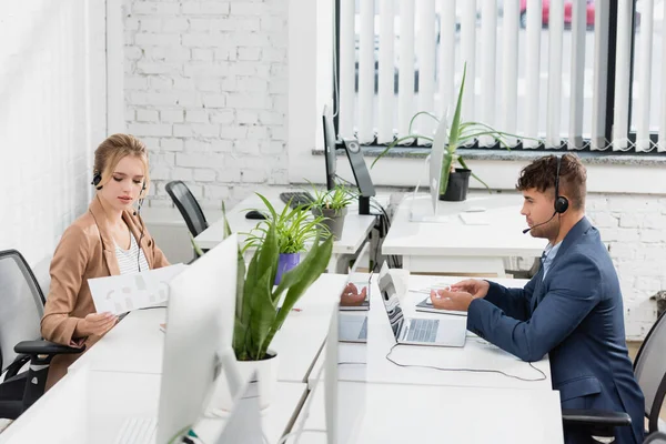 Operatori di call center in cuffie funzionanti, seduti ai tavoli con dispositivi digitali in ufficio — Foto stock