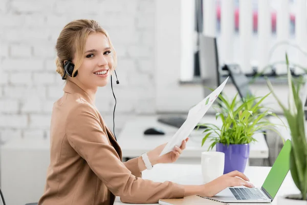 Femme souriante dans un casque avec feuille de papier regardant la caméra, tout en étant assis sur le lieu de travail avec un ordinateur portable sur fond flou — Photo de stock