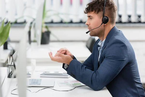 Vue latérale de l'homme en gestuelle casque, tout en regardant l'ordinateur portable sur le lieu de travail sur le premier plan flou — Photo de stock