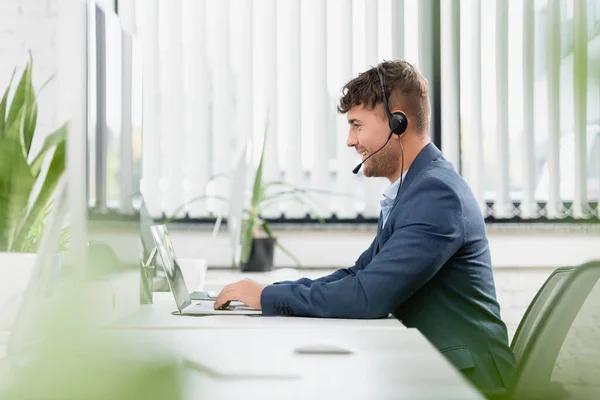 Lächelnder Mann mit Headset tippt auf Laptop, während er am Arbeitsplatz sitzt, mit Pflanzen im Büro auf verschwommenem Vordergrund — Stockfoto