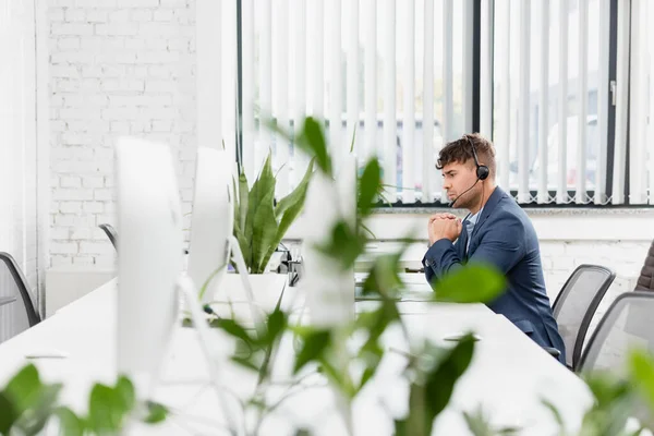 Operador sério no fone de ouvido sentado no local de trabalho no escritório com a planta turva em primeiro plano — Fotografia de Stock