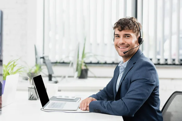 Fröhlicher Berater im Headset blickt in die Kamera, während er mit digitalen Geräten auf verschwommenem Hintergrund am Tisch sitzt — Stockfoto