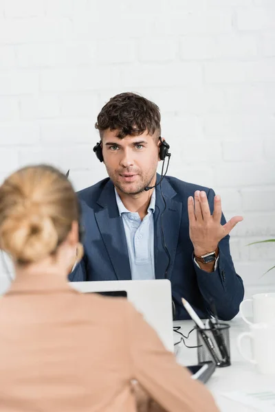 Vista posteriore della donna seduta vicino all'operatore in cuffia gesticolare sul posto di lavoro con dispositivi digitali in primo piano sfocati — Foto stock