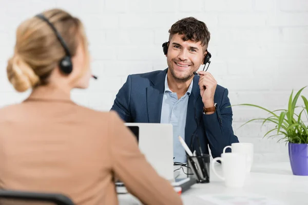 Smiling operator in headset looking at camera, while sitting at workplace with blurred colleague on foreground — Stock Photo