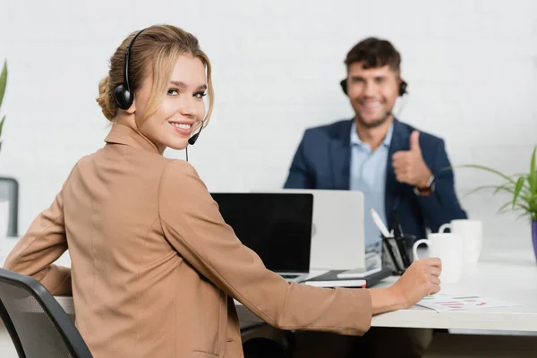 Donna sorridente in auricolare guardando la fotocamera, mentre seduto sul posto di lavoro con collega offuscata sullo sfondo — Foto stock