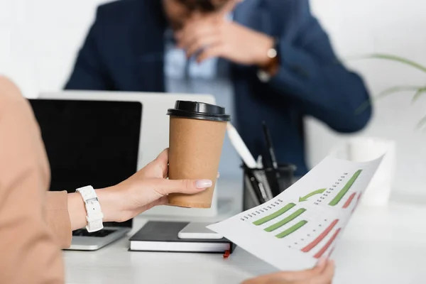 Cropped view of businesswoman with paper cup, holding paper sheet with graphs at work on blurred background — Stock Photo