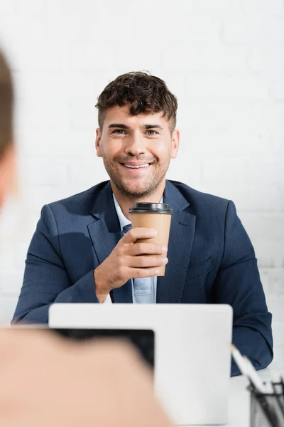 Homme d'affaires souriant avec tasse en papier regardant son collègue, assis sur le lieu de travail au premier plan flou — Photo de stock