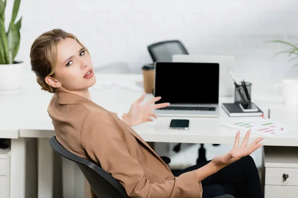 Mujer ejecutiva irritada mirando la cámara y el gesto, mientras está sentada en el lugar de trabajo con dispositivos digitales sobre un fondo borroso — Stock Photo