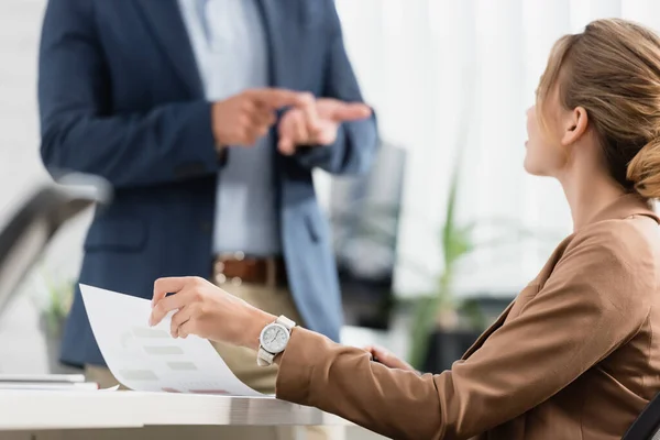 Female executive with document sitting at table with blurred colleague gesturing on background — Stock Photo