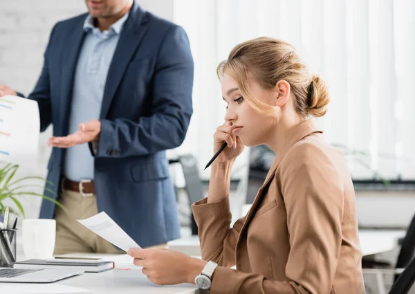Aufgebrachte Geschäftsfrau mit Stift schaut auf Dokument, während sie am Arbeitsplatz mit verschwommenem Kollegen im Hintergrund sitzt — Stockfoto