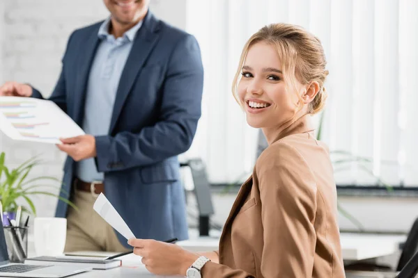 Happy businesswoman with document looking at camera, while sitting at workplace with blurred colleague on background — Stock Photo
