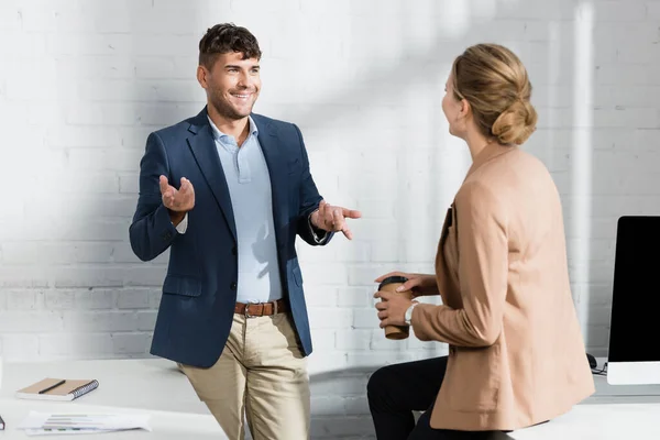 Smiling businessman gesturing, while talking to colleague near workplace during break in office — Stock Photo