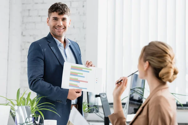 Lächelnder Geschäftsmann blickt in die Kamera, während er Papierblätter mit Diagrammen in der Nähe seines Arbeitsplatzes im unscharfen Vordergrund zeigt — Stockfoto