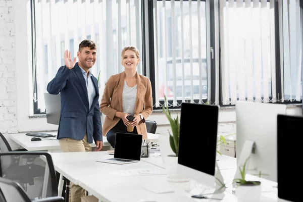 Feliz hombre de negocios con la mano agitada, mirando a la cámara, mientras está de pie cerca de su compañero de trabajo durante el descanso en la oficina - foto de stock