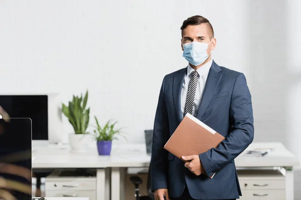 Empresario en carpeta de retención de máscara médica, mientras está de pie en la oficina sobre fondo borroso - foto de stock