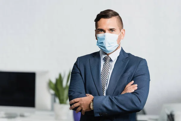 Businessman in medical mask with crossed arms, looking at camera with blurred office on background — Stock Photo