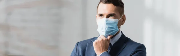 Thoughtful businessman in medical mask looking away on blurred background, banner — Stock Photo